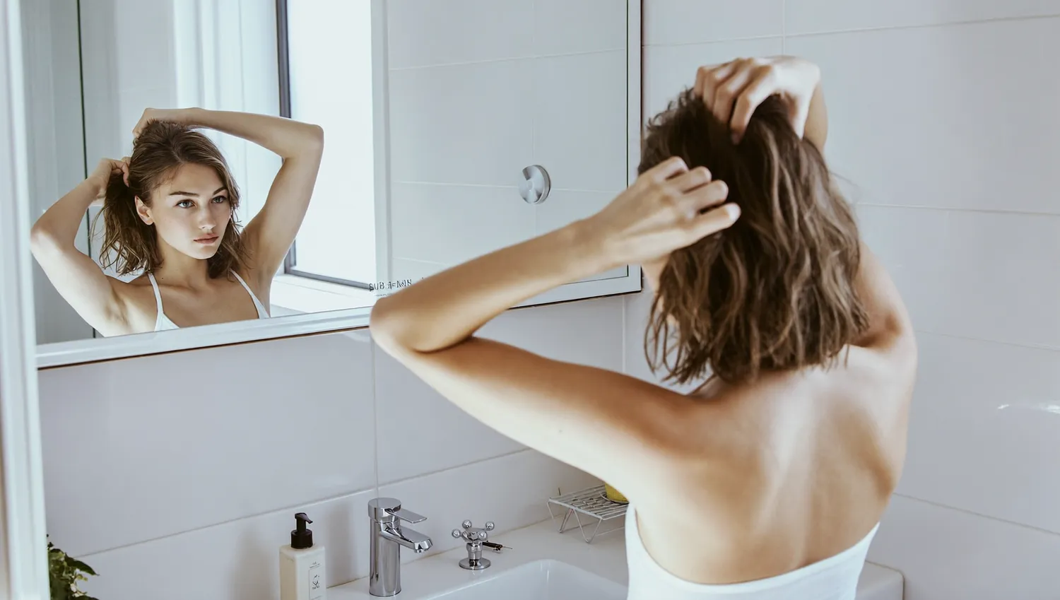洗面所の鏡の前で、頭皮に触れながら自分の髪を気にかける女性の後ろ姿