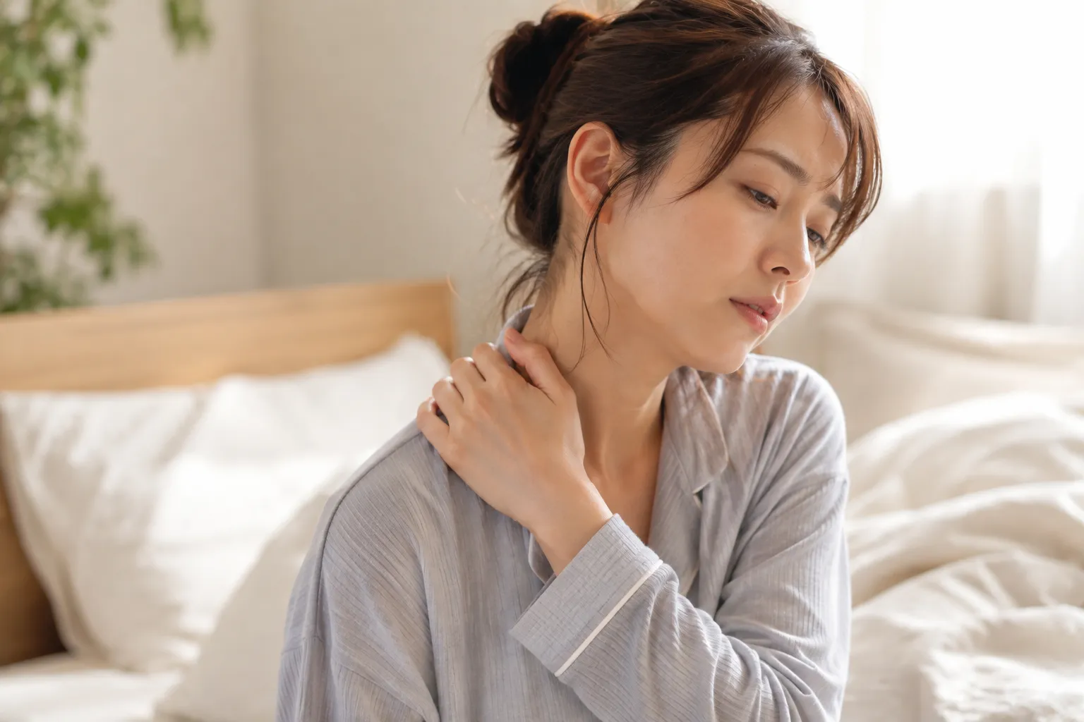 ベッドの上で自分の肩・首をさすり、不調そうな表情を見せる女性