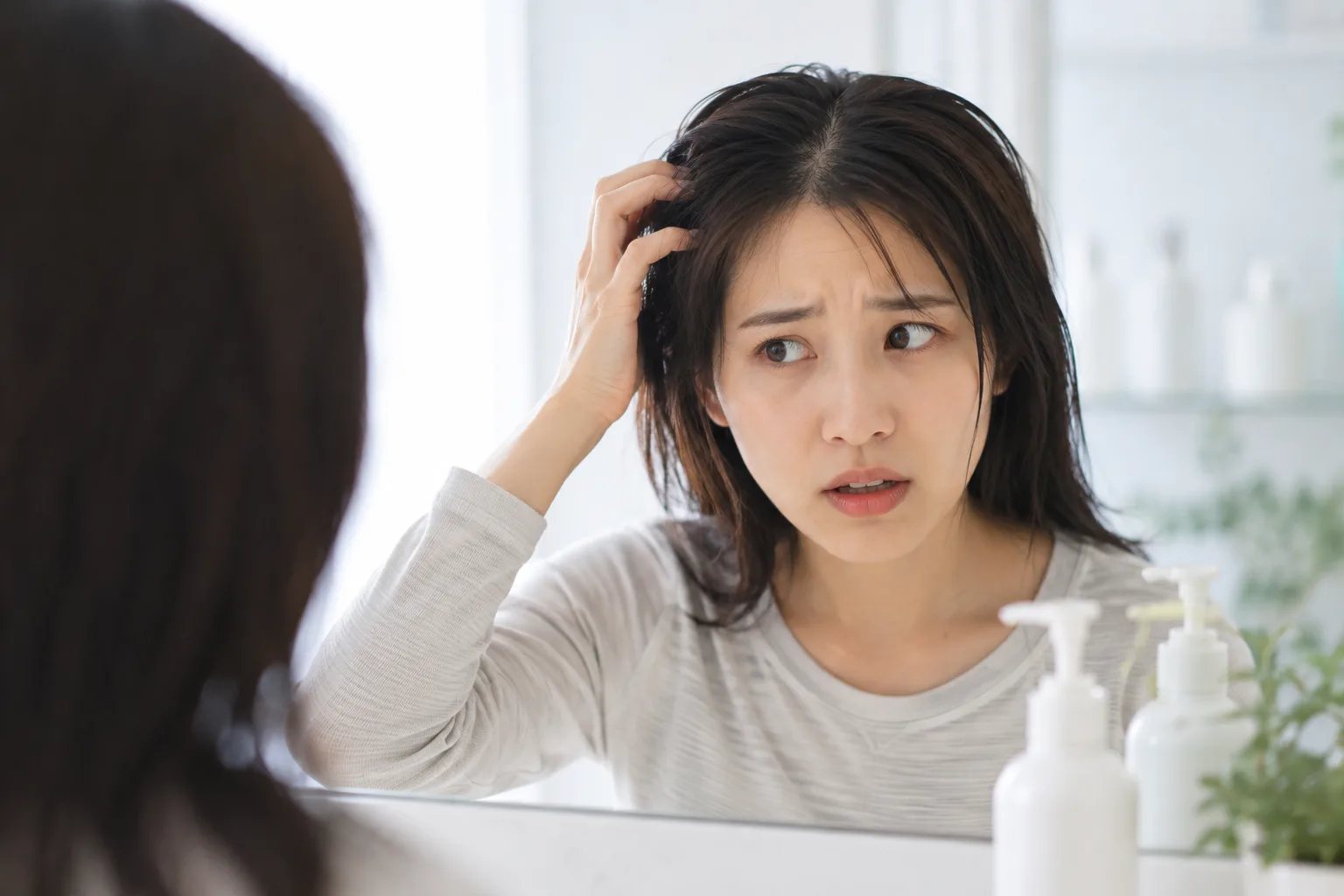 鏡の前で頭皮に手をあて、ピリピリとした違和感に驚き不安げな表情を見せる女性